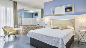 Una habitación de hotel moderna con una cama grande, un área de descanso con una silla y una mesita, un baño con ducha y espejo, y un ventanal con cortinas.