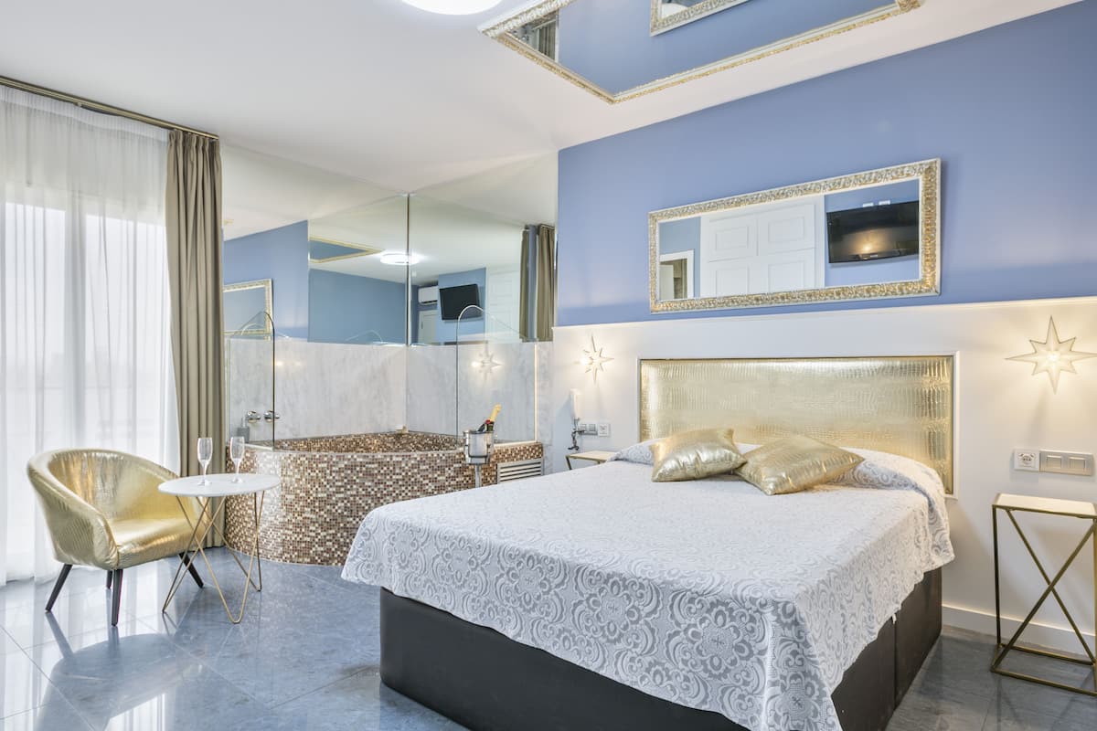 Una habitación de hotel moderna con una cama grande, un área de descanso con una silla y una mesita, un baño con ducha y espejo, y un ventanal con cortinas.