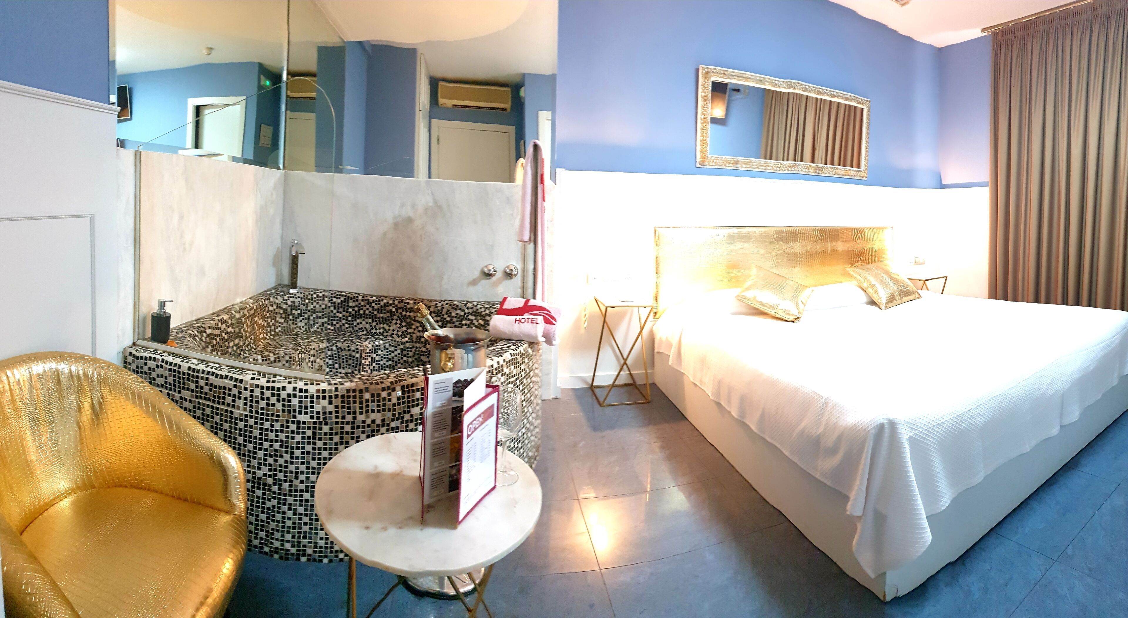 Habitación de hotel con una cama grande, una zona de estar con una silla y una mesita, una bañera con mosaicos y un espejo grande en la pared.