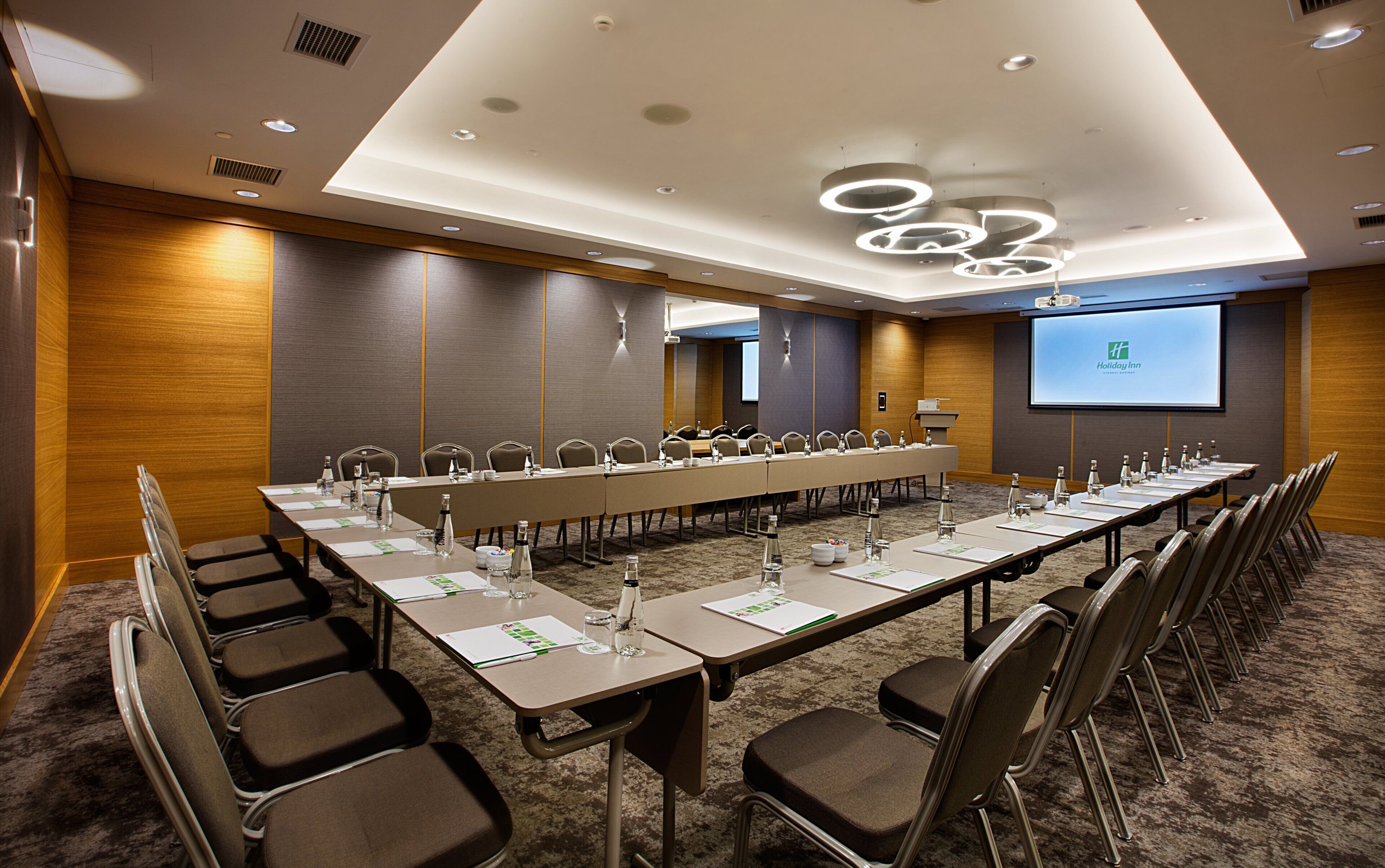Un salón de conferencias con una larga mesa, sillas y una pantalla que muestra el logotipo de Holiday Inn.