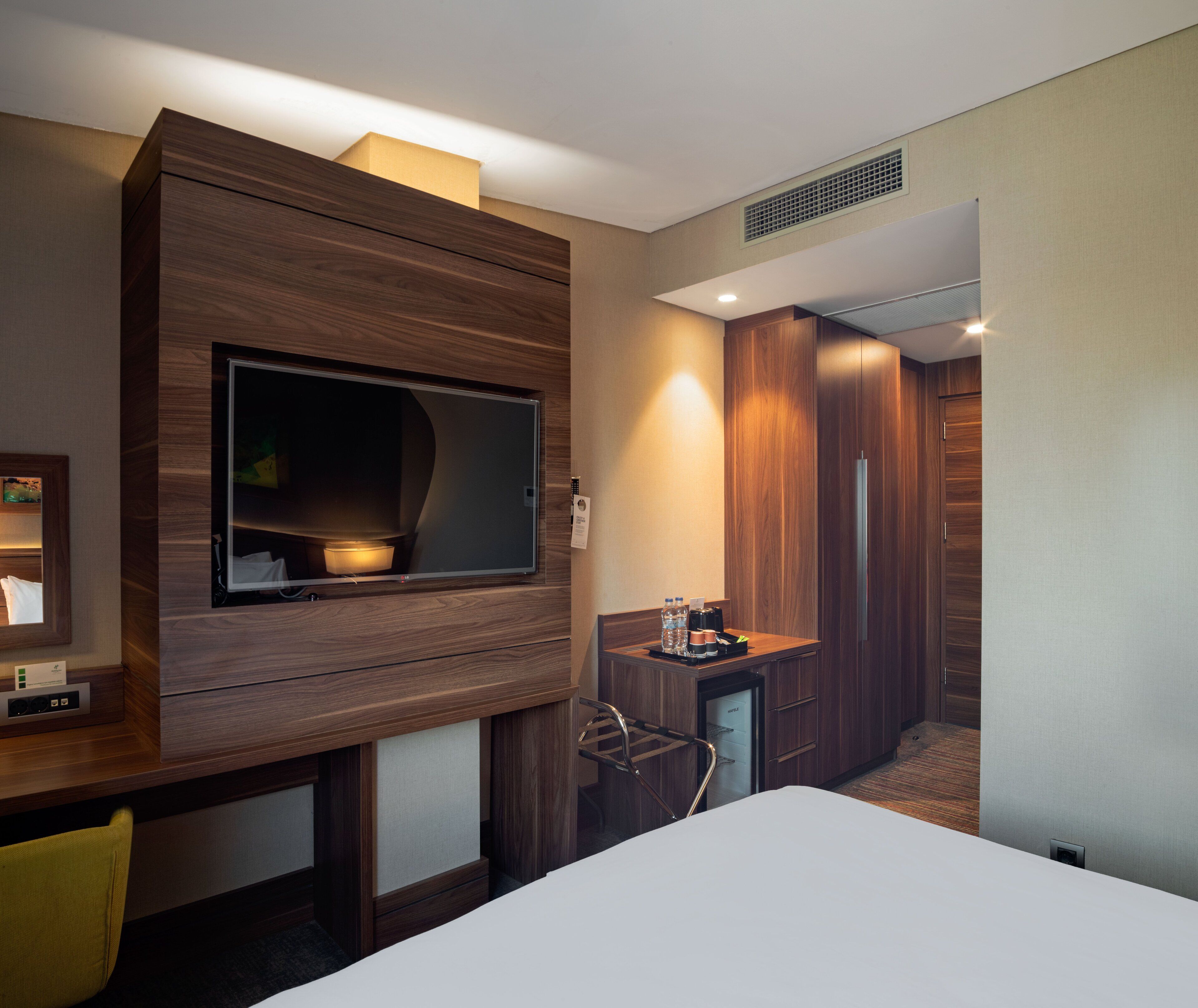 A hotel room with a large flat-screen TV mounted on a wooden wall, a small desk with a chair, a minibar, and a separate bathroom area.