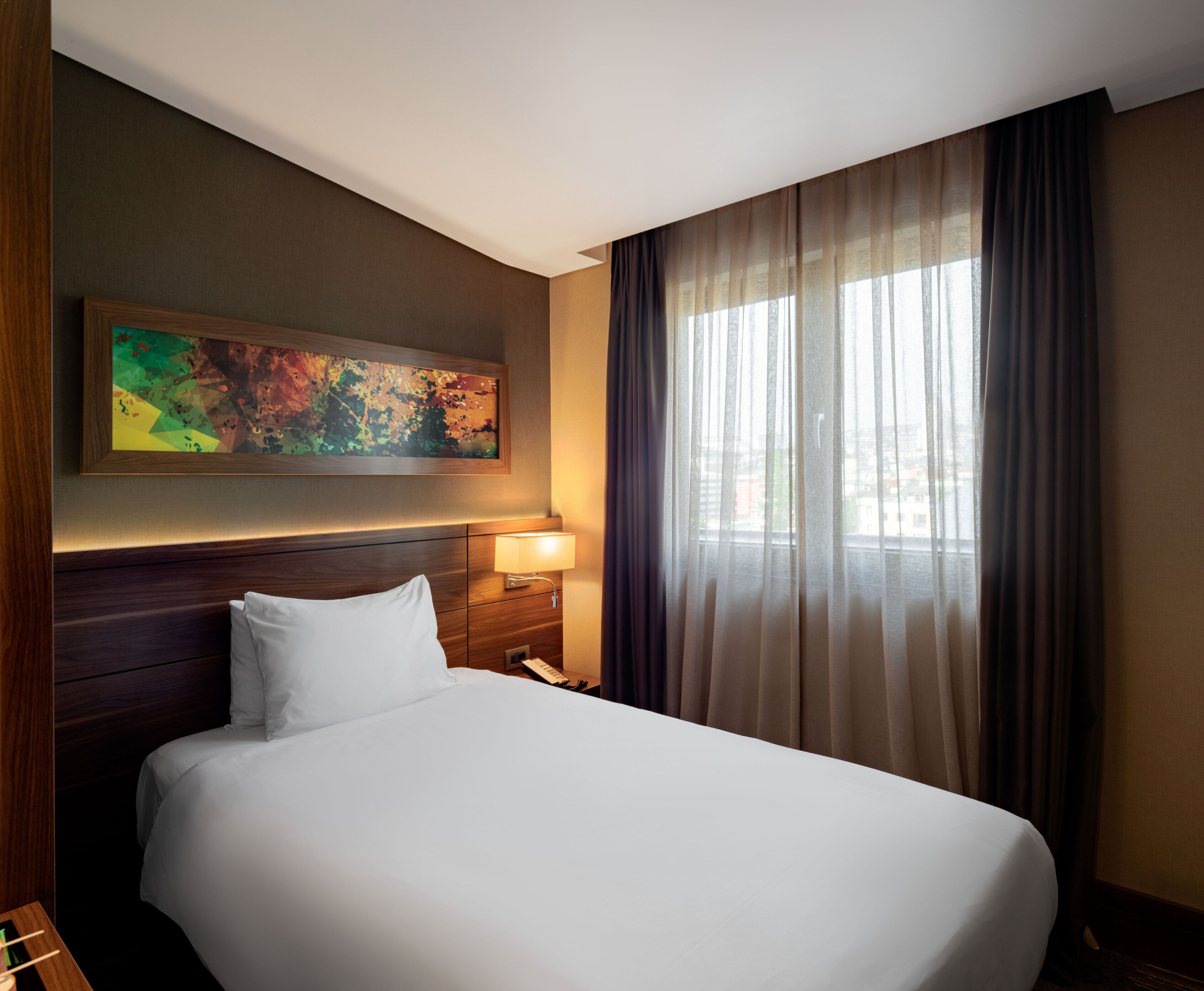 A hotel room with a large bed, a nightstand with a lamp, a painting on the wall, and a window with curtains.