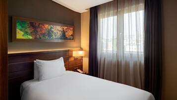 A hotel room with a large bed, a nightstand with a lamp, a painting on the wall, and a window with curtains.