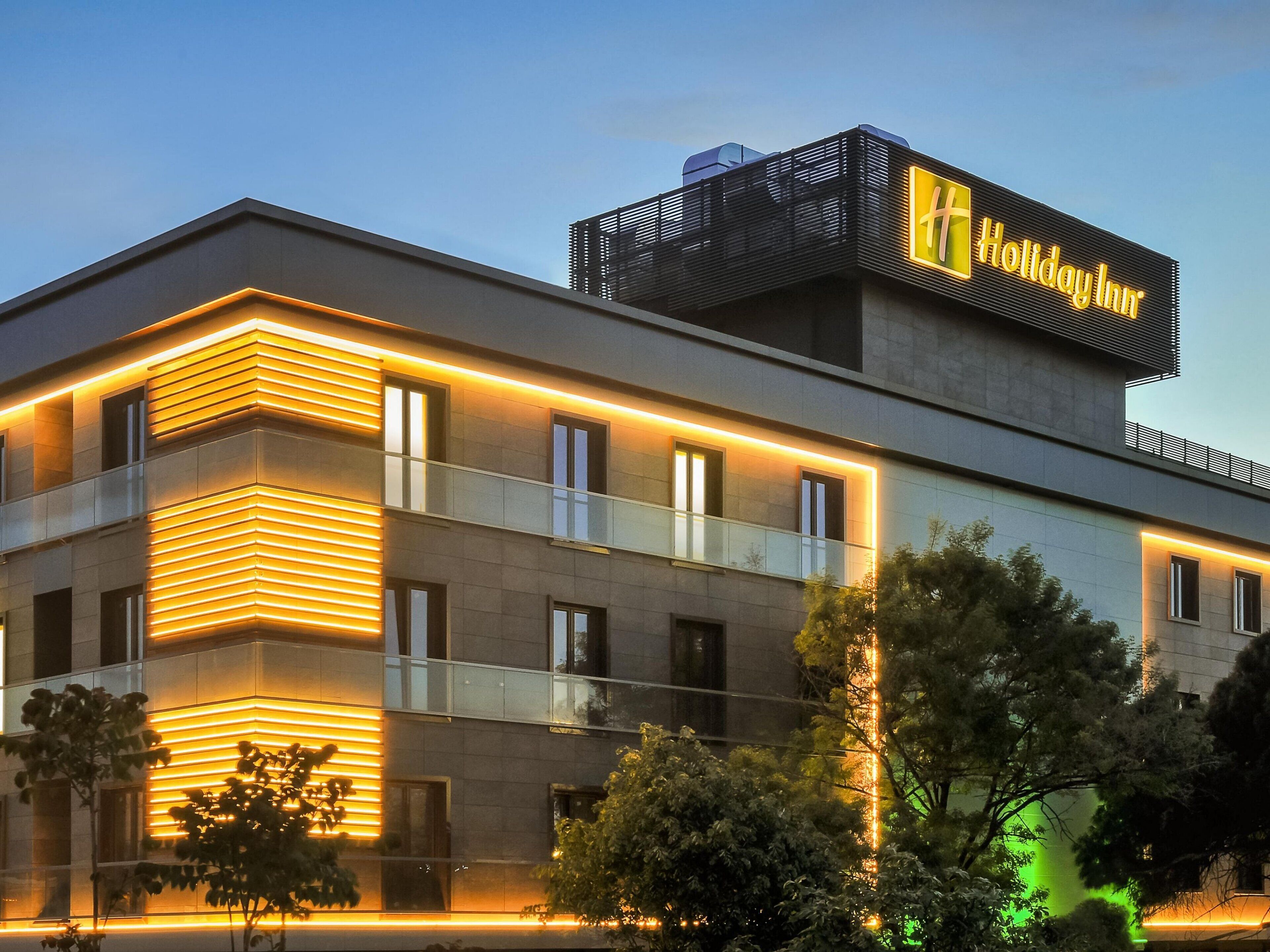 Un edificio moderno con un llamativo letrero de 'Holiday Inn', con techo plano, ventanas amplias y un balcón con barandillas de vidrio.