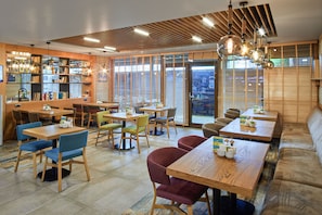 Un café moderne avec des tables et des chaises en bois, une bibliothèque et de grandes fenêtres équipées de stores.