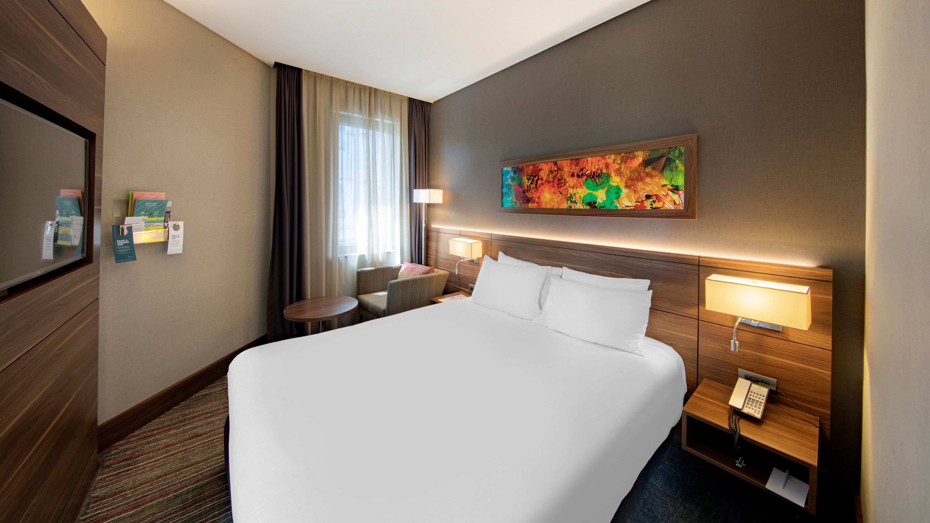A hotel room with a large bed, a desk, a chair, a TV, and a painting on the wall.