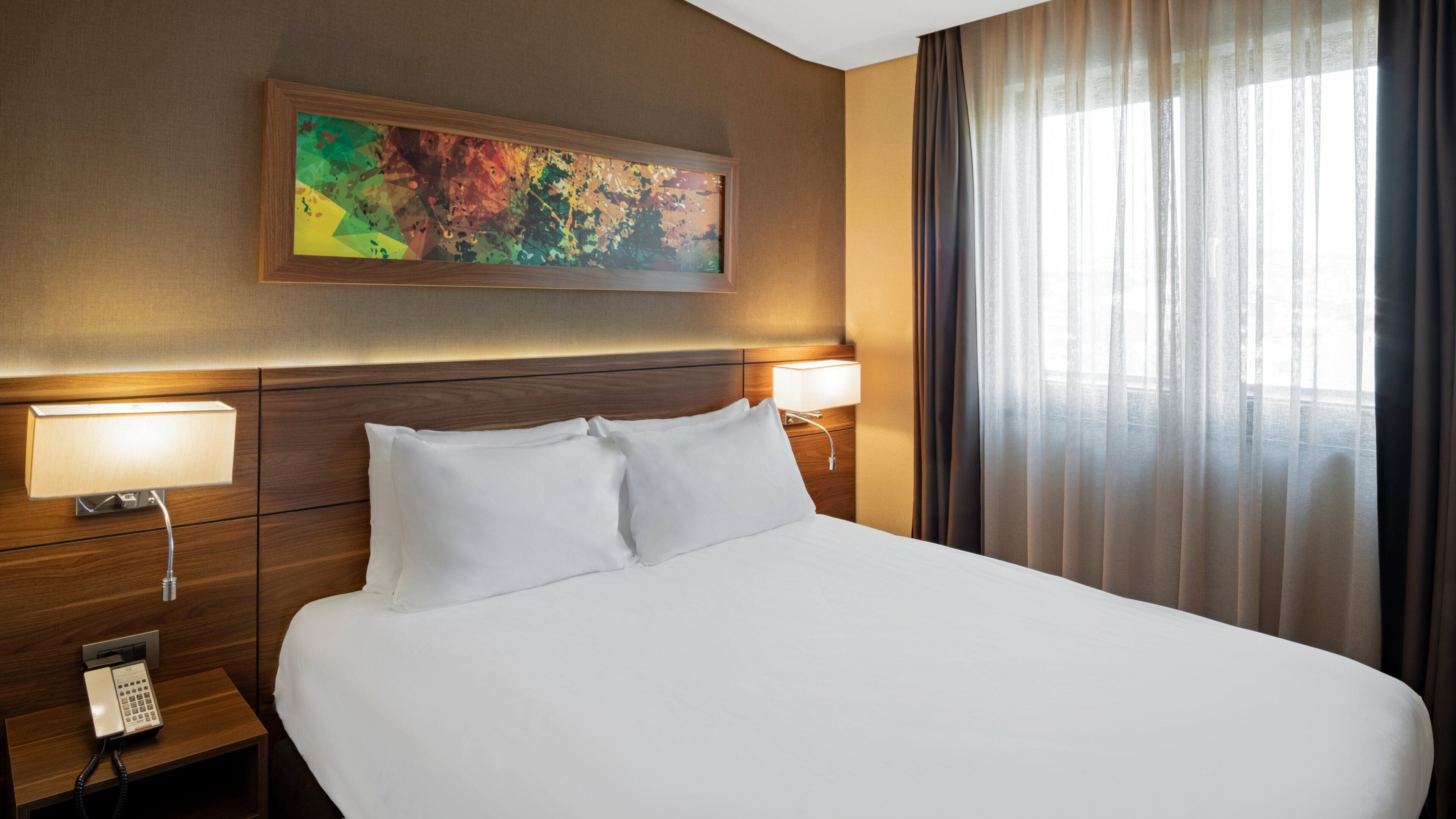 Una habitación de hotel con una cama grande, dos lámparas de noche, un teléfono y un cuadro abstracto y colorido en la pared.