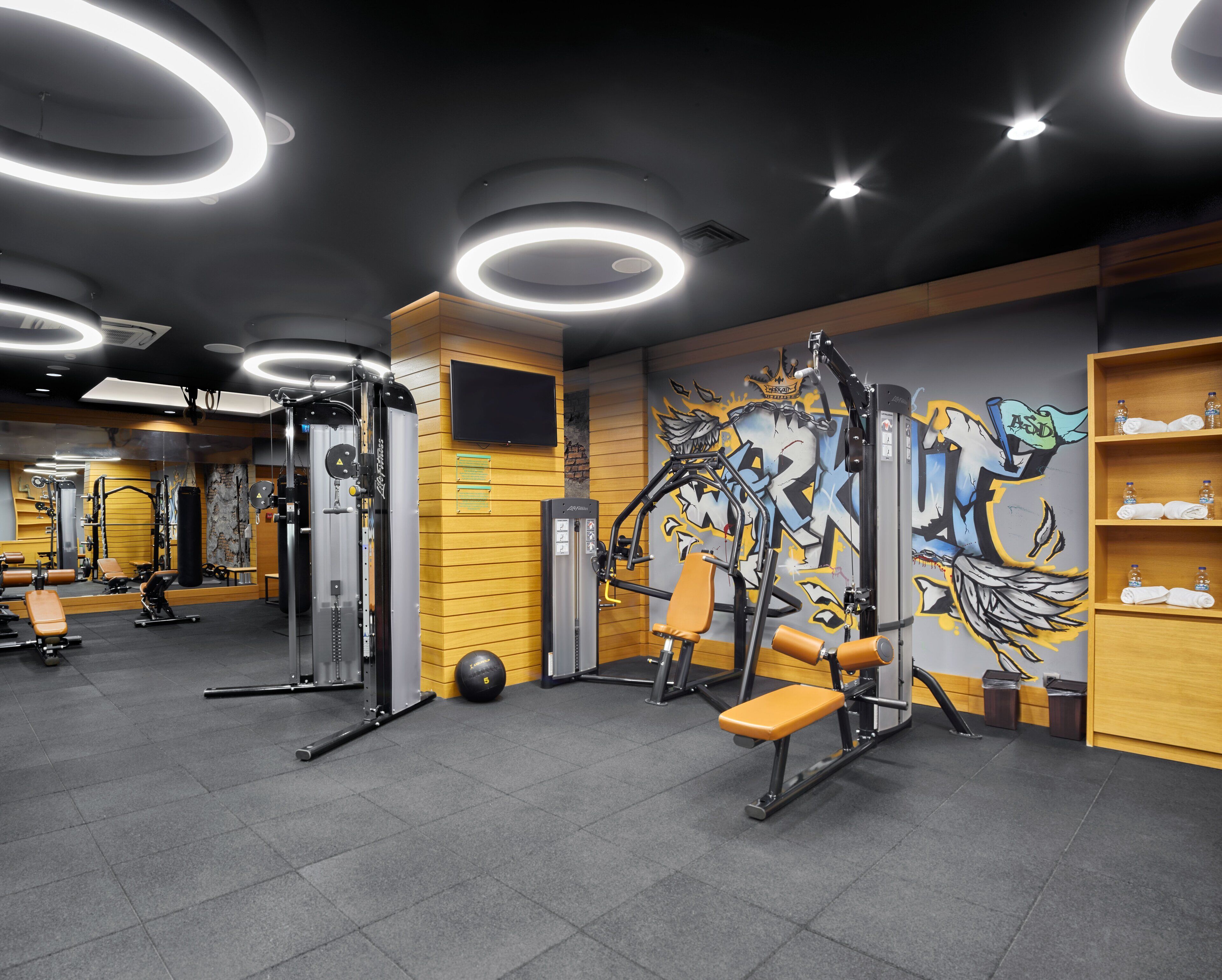 Uma academia com equipamentos de ginástica, espelho e um muro com grafites.