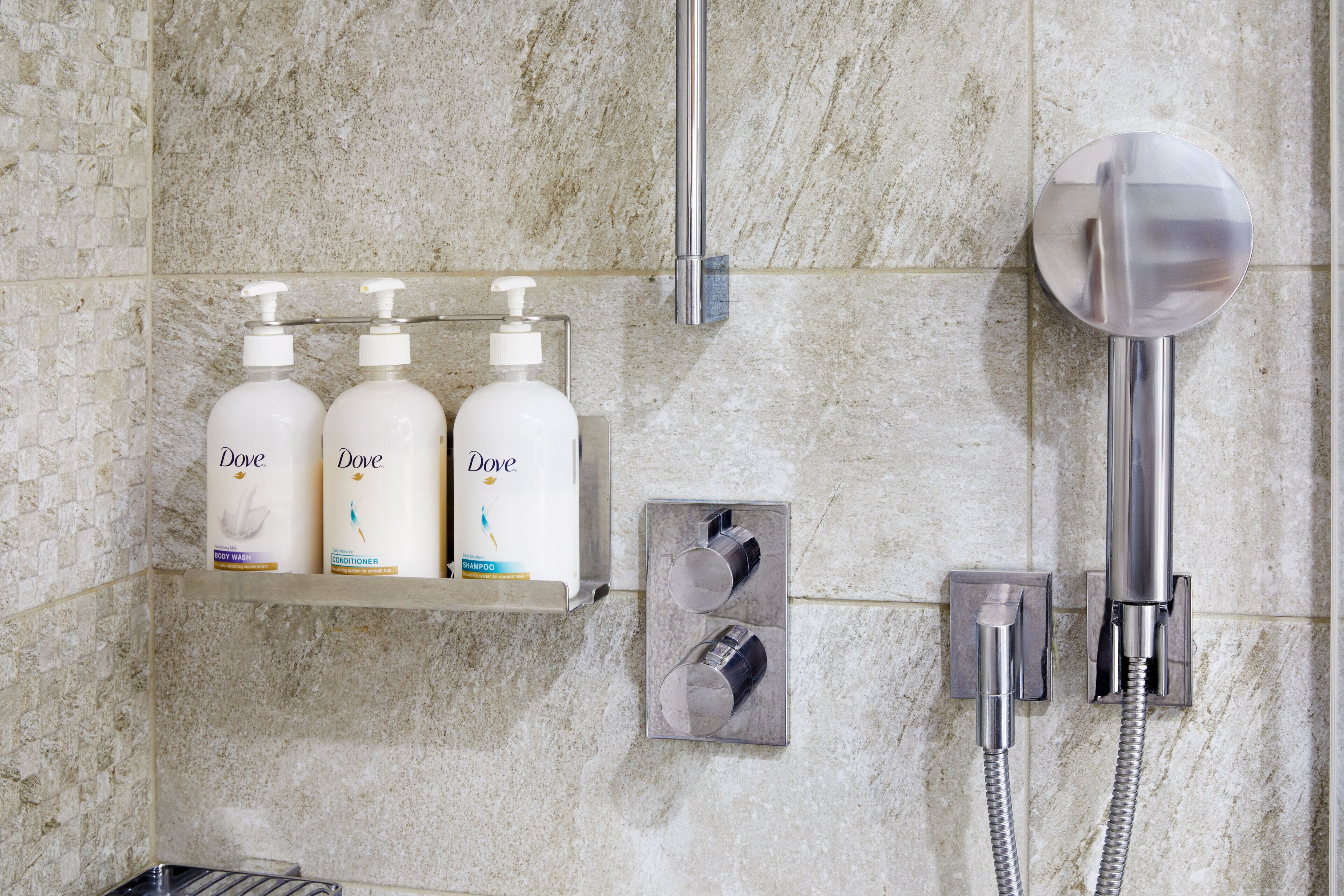 A bathroom with a marble wall, a shelf with Dove body wash, conditioner, and shampoo, a handheld showerhead, and a towel rack.