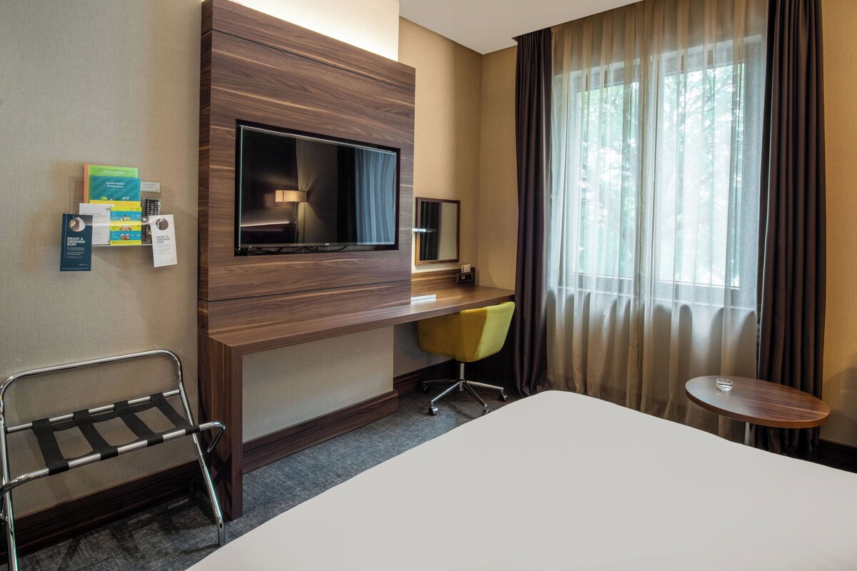 Een hotelkamer met een bed, een bureau met televisie, een stoel, een klein tafeltje en een raam met gordijnen.
