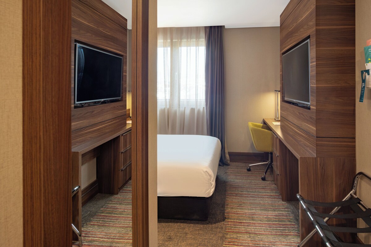 Hotelkamer met een bed, twee televisies, een bureau, een stoel en een raam met gordijnen.