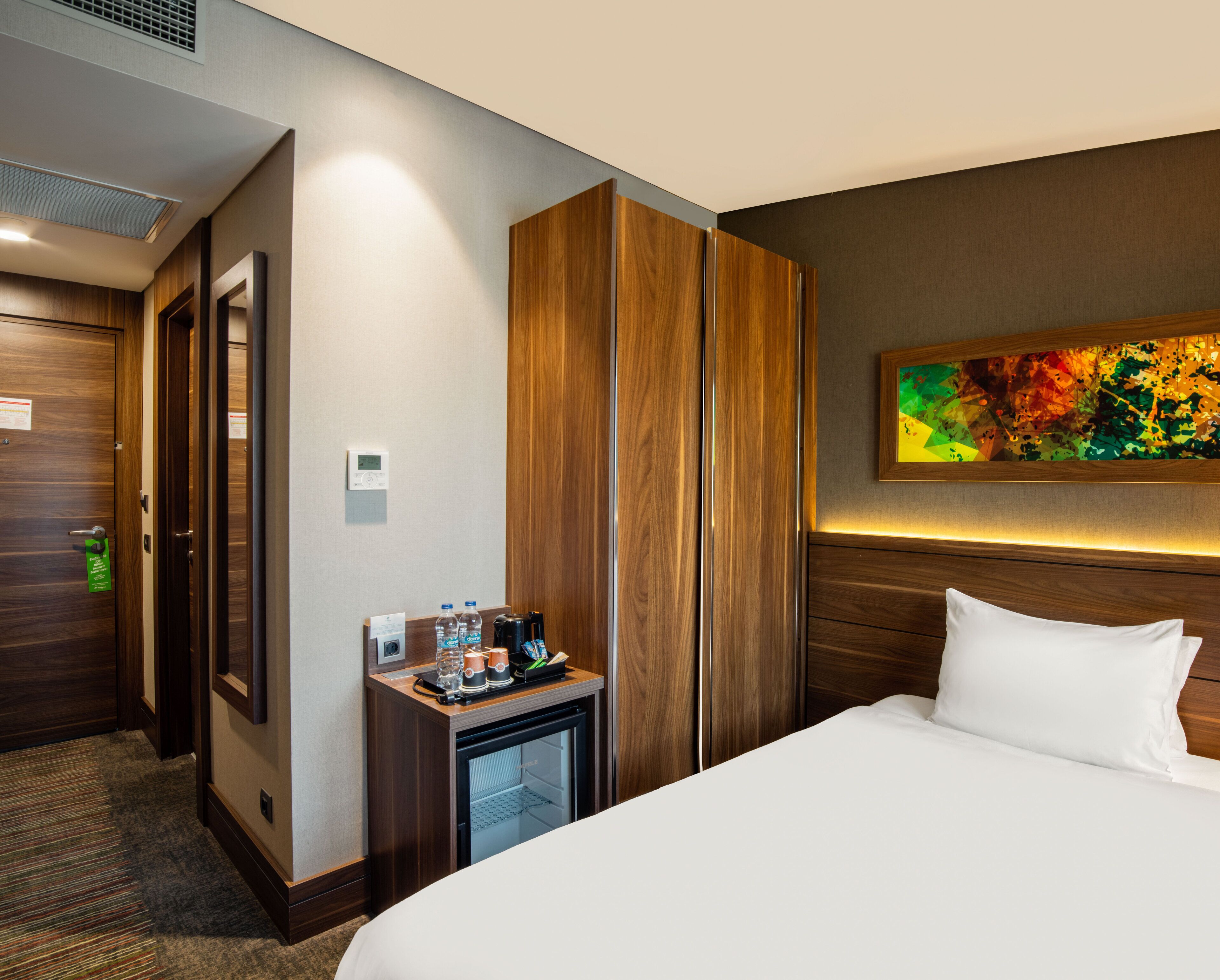 Una habitación de hotel con cama, un mini bar, un armario de madera, un cuadro en la pared y un termostato.