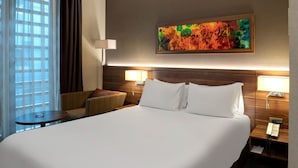 Hotellihuone, jossa on suuri sänky, kaksi yöpöytää valaisimineen, tuoli ja ikkuna, jossa on verhot.