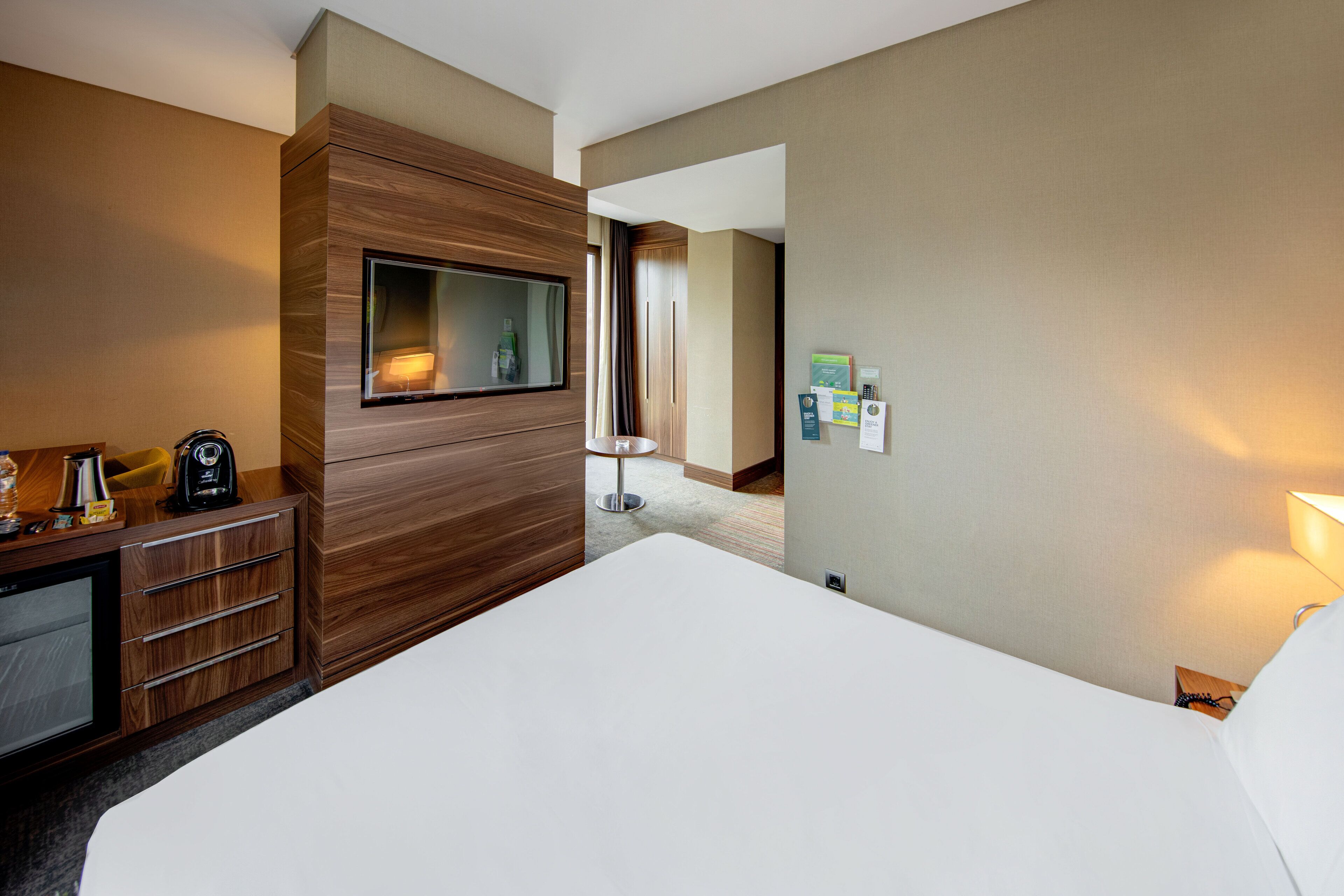 A hotel room with a large bed, a wooden panel wall, a TV, a desk with a lamp, and a small table with a bottle.