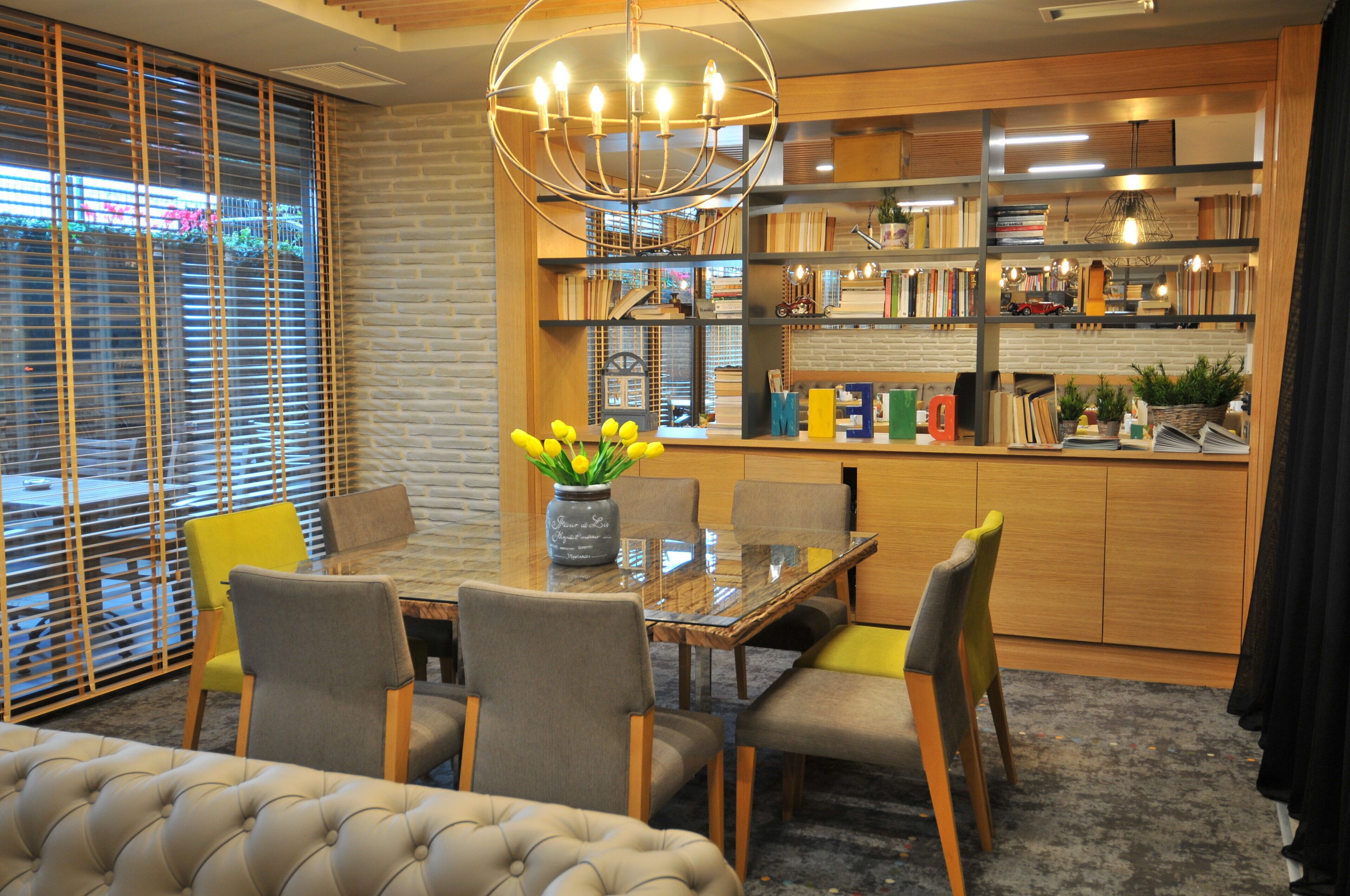 A dining area with a glass table, four chairs, a vase with yellow flowers, and a bookshelf filled with books and decorative items.