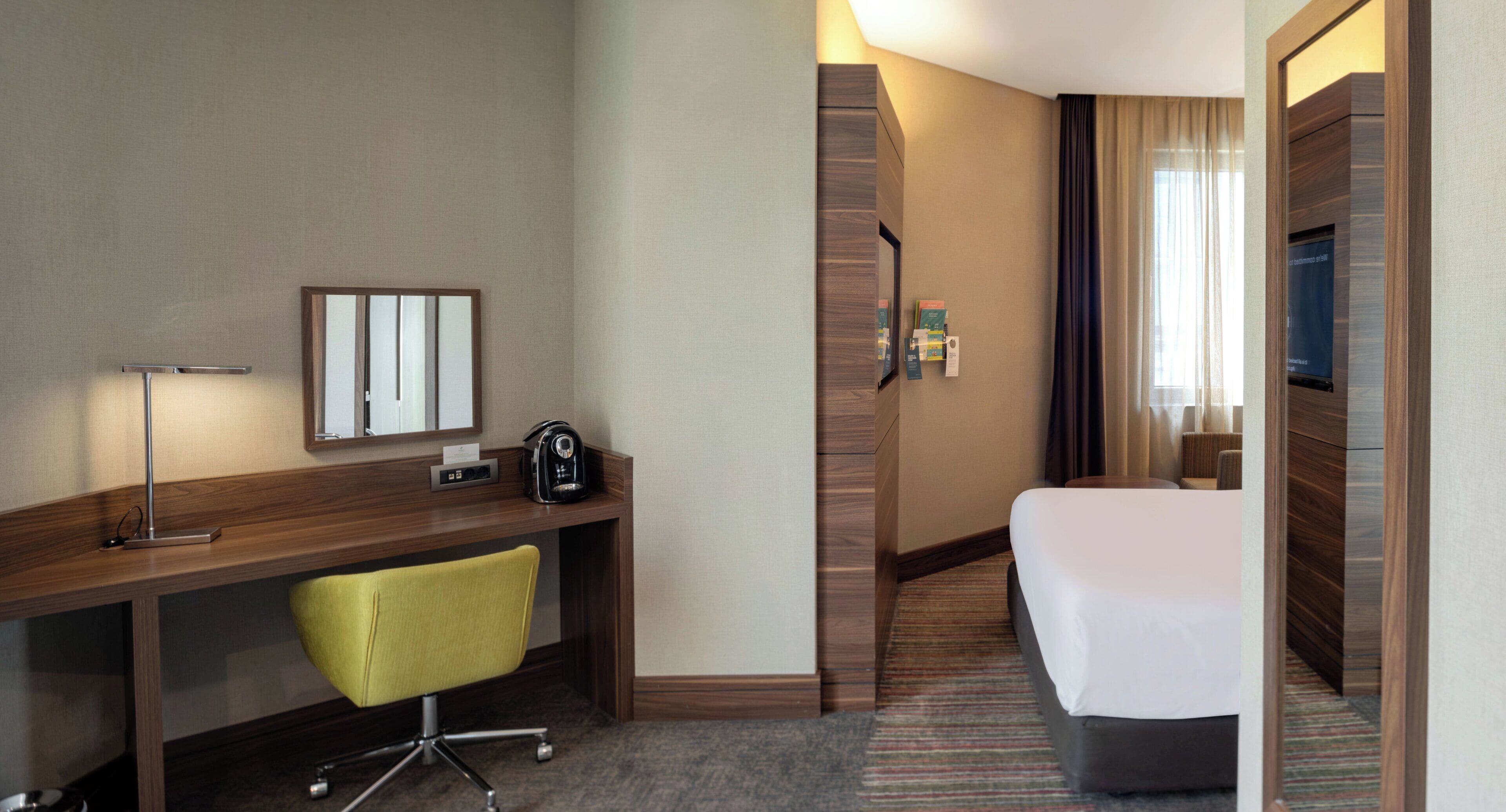 A hotel room with a wooden desk, a yellow chair, a bed, a TV, and a mirror.