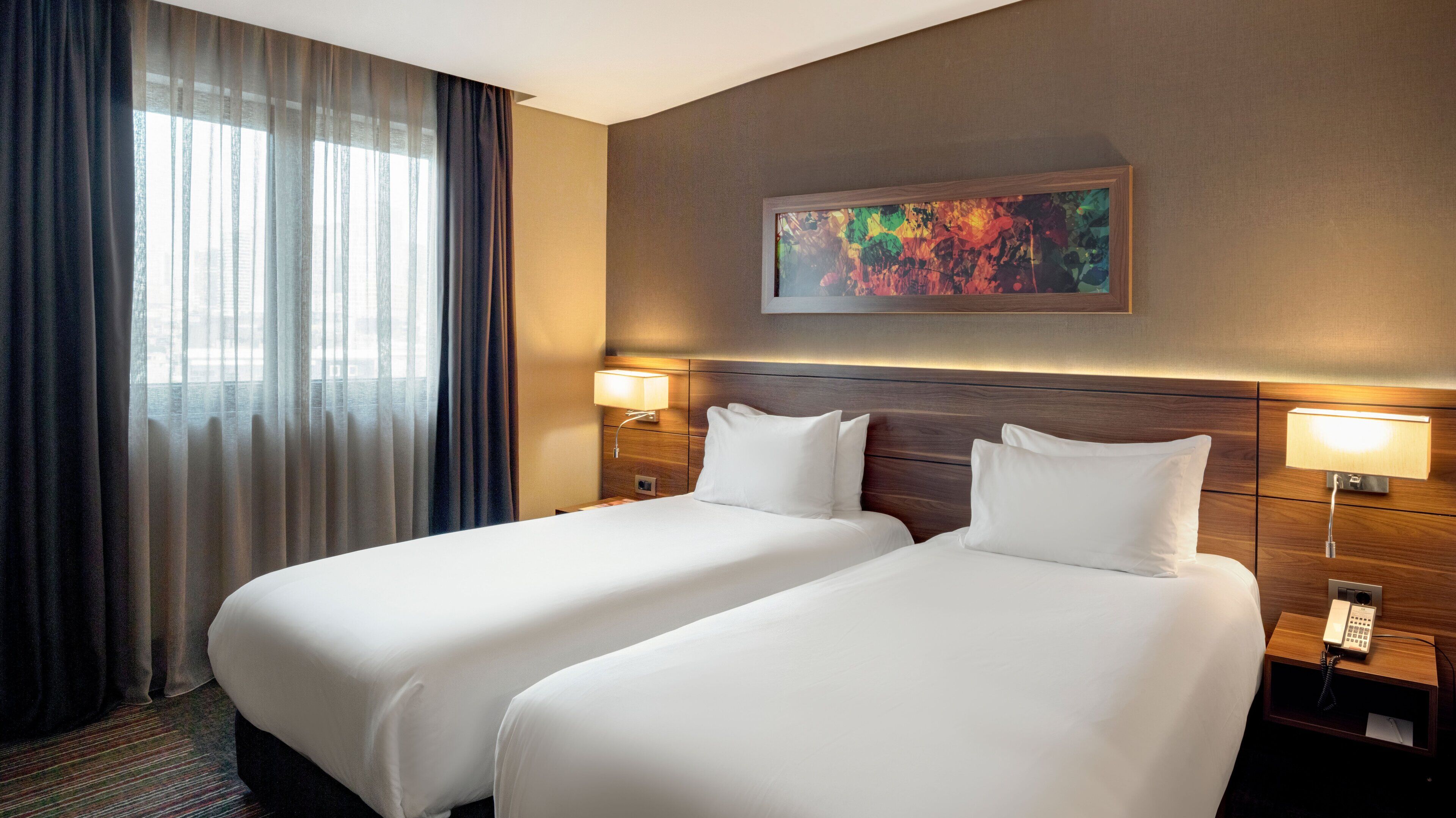 A hotel room with two beds, a large window with sheer curtains, a painting on the wall, and bedside lamps.
