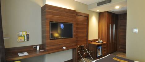 A hotel room with a flat-screen TV mounted on a wooden panel, a desk with a remote control, a chair, a bed, and a minibar.