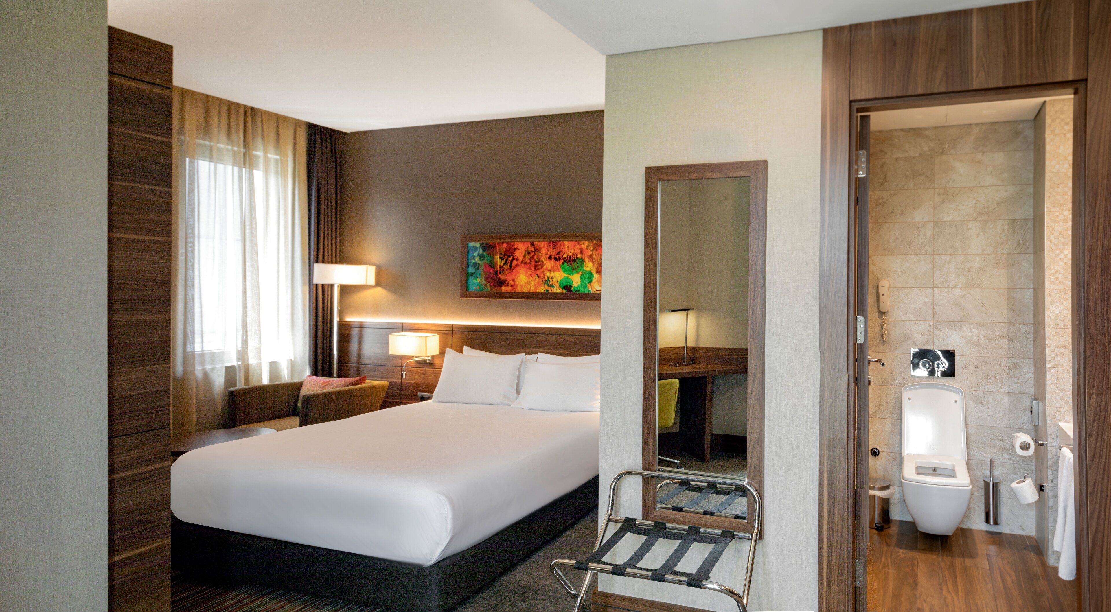 A hotel room with a large bed, a bedside table with a lamp, a small armchair, a bathroom with a toilet and sink, and a shower area.
