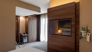 Moderni hotellihuone, jossa on suuri televisio, minibaari ja työpöytä tuolilla.