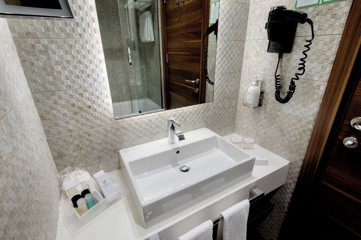 Een badkamer met een witte wastafel, een douchecabine van glas, een houten deur en een aan de muur bevestigd telefoon.