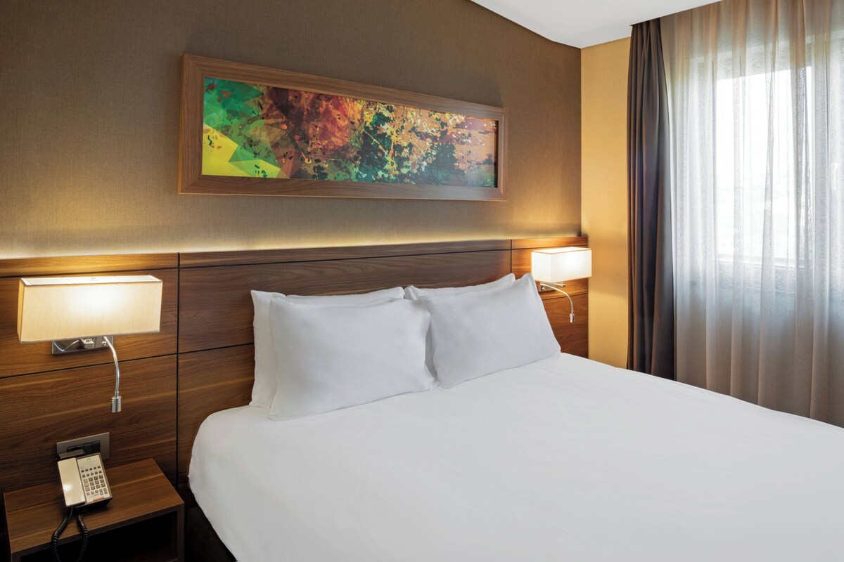 Hotelkamer met een groot bed, twee bedlampen, een nachtkastje met telefoon, een schilderij aan de muur en een raam met gordijnen.