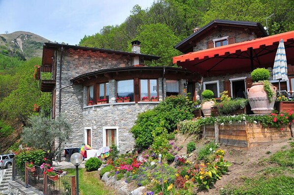 A stone house with a balcony, surrounded by colorful flowers and plants. - Agriturismo La Fonte di Mariella (Dosso del Liro)