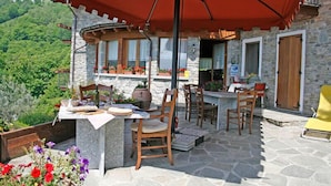 Eine Steinterrasse mit Gartenmöbeln, einem Tisch mit rotem Sonnenschirm und Blick auf einen üppigen, grünen Hang.