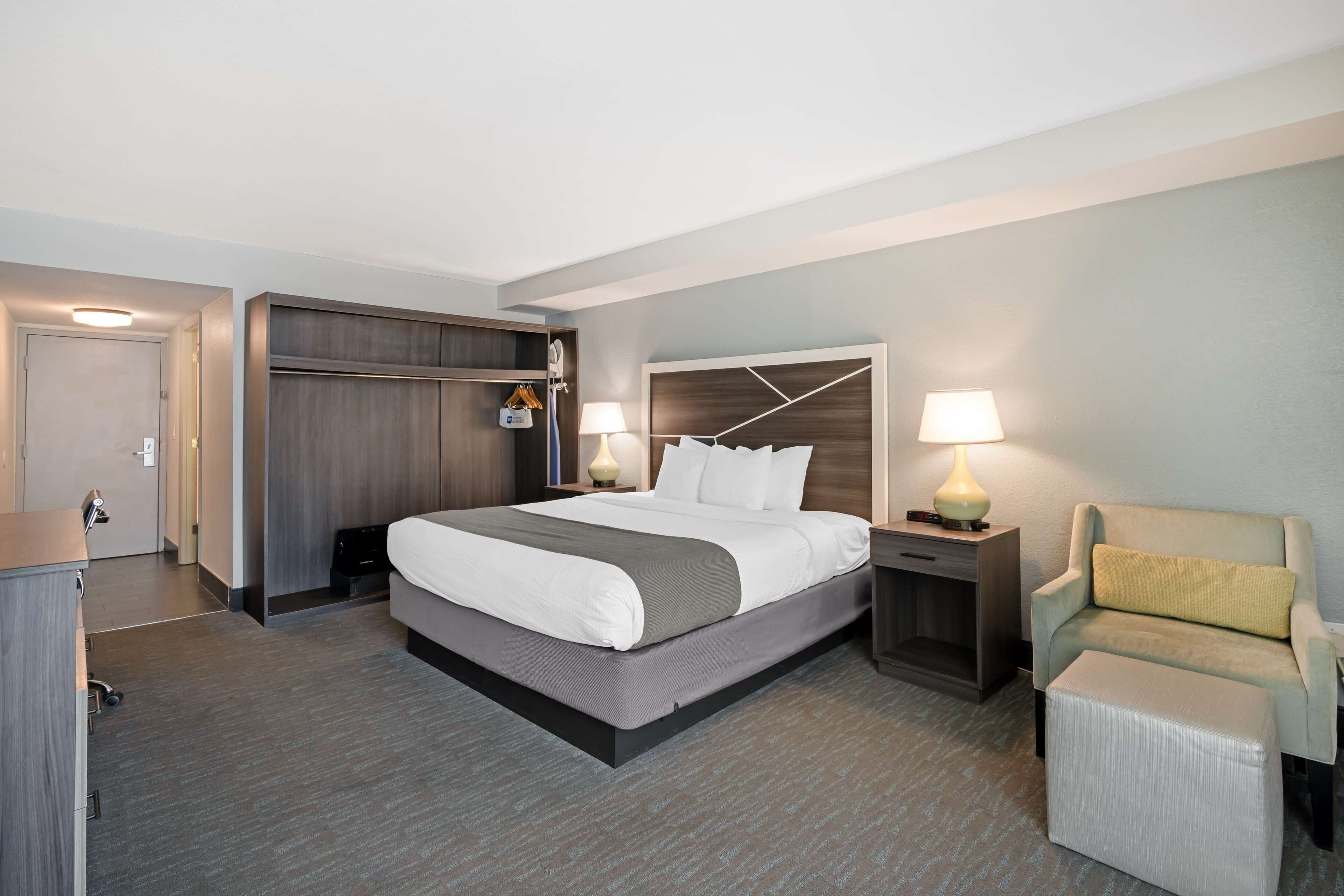 A modern hotel room with a large bed, a desk, a chair, and a wardrobe.