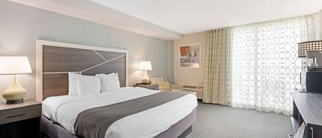 A hotel room with a large bed, two bedside tables with lamps, a desk with a chair, a window with patterned curtains, and a wall-mounted TV.