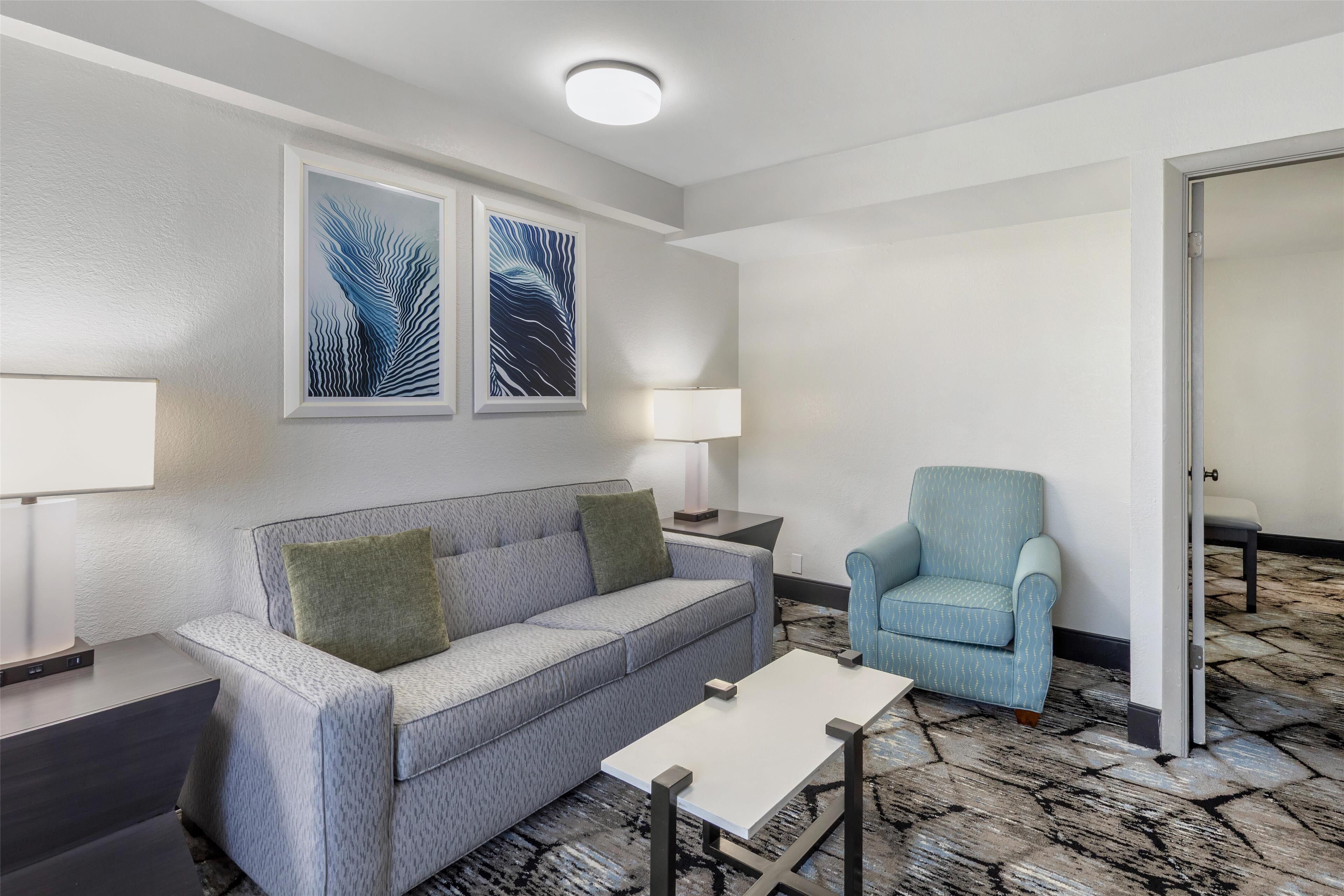 A modern hotel room with a grey sofa, a blue armchair, a white table, and two framed artworks on the wall.