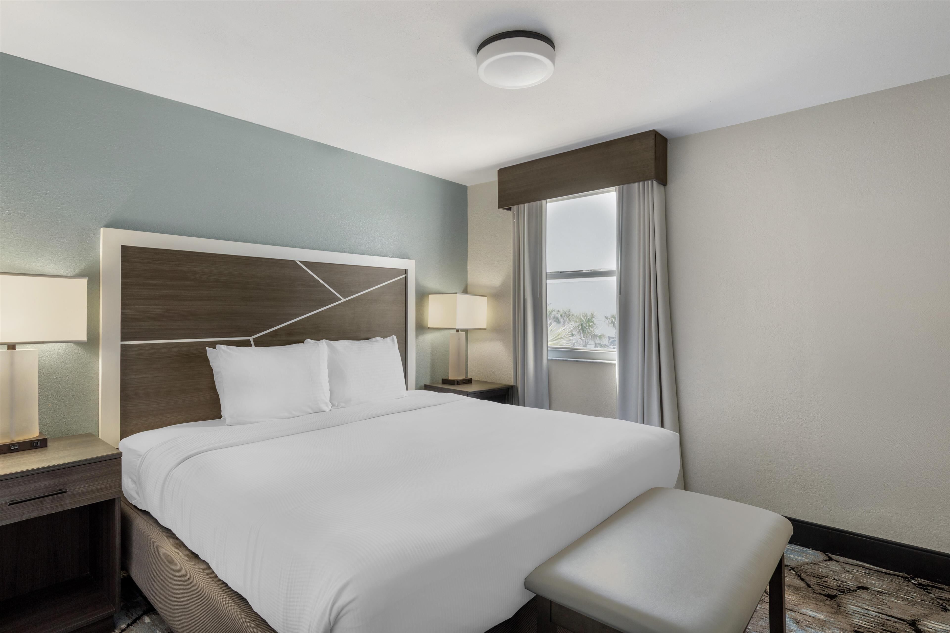 A hotel room with a large bed, two bedside tables with lamps, a bench, and a view of the outdoors through a window.