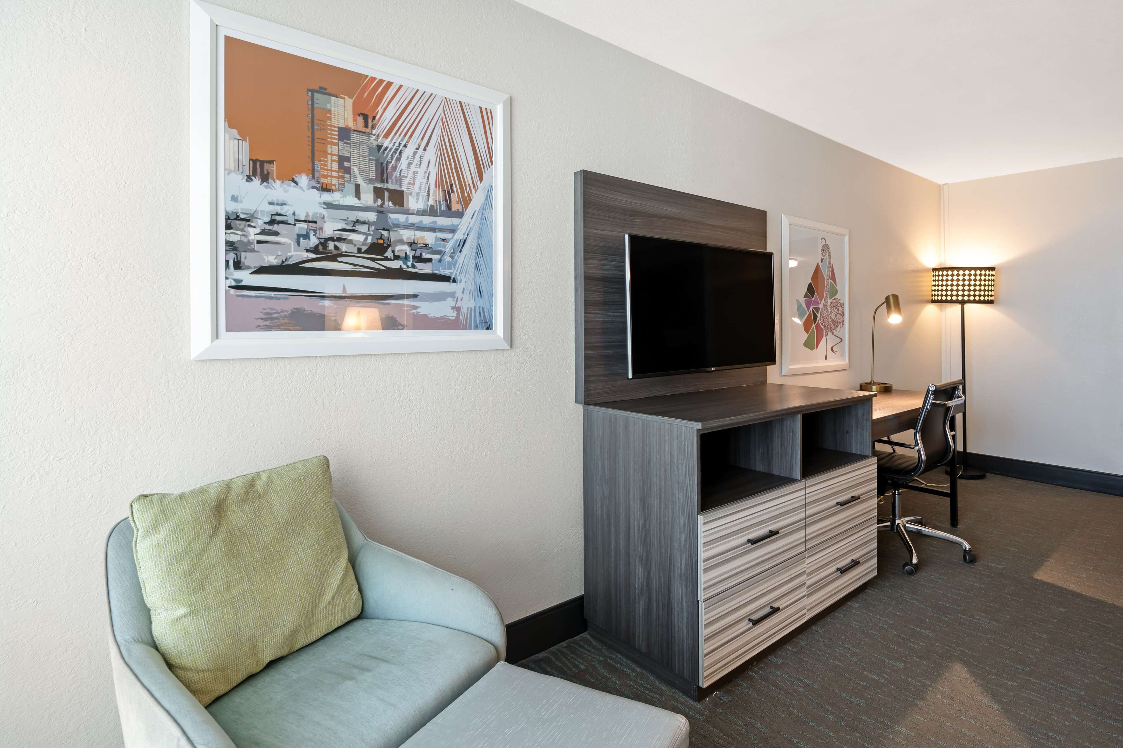 Habitación de hotel con sofá, televisión montada en un mueble de madera, escritorio con silla y dos cuadros en la pared.