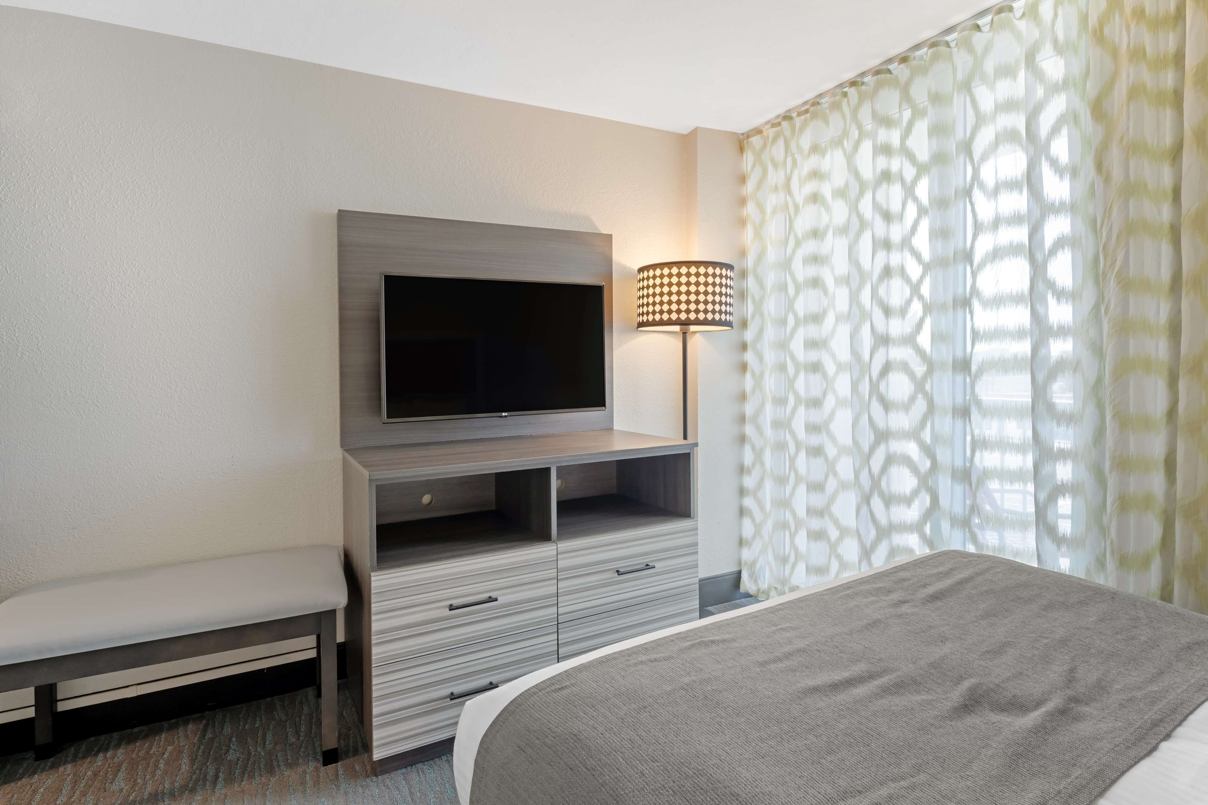 A modern hotel room with a flat-screen TV mounted on a wooden cabinet, a bedside lamp, a bench, and a large bed.
