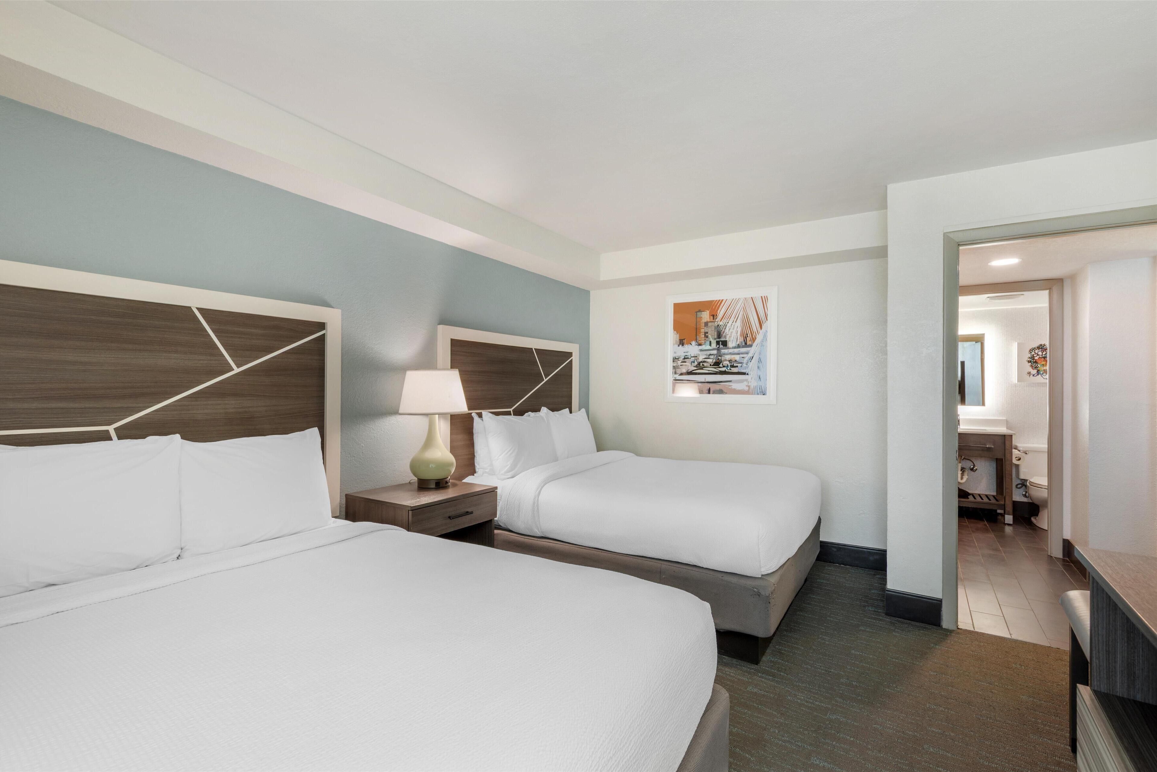 A hotel room with two beds, a nightstand with a lamp, a painting on the wall, and a bathroom visible through an open door.