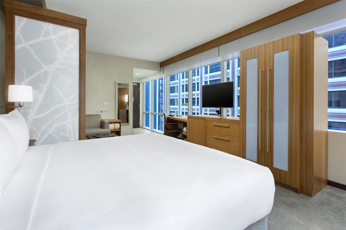 A hotel room with a large bed, wooden wardrobe, a desk with a chair, a television, and large windows offering a view of the city.