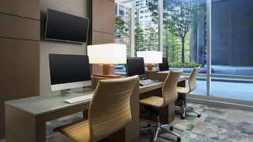 A modern office space with two desks, two chairs, a flat-screen monitor, a desktop computer, and a large window offering a view of buildings and trees.