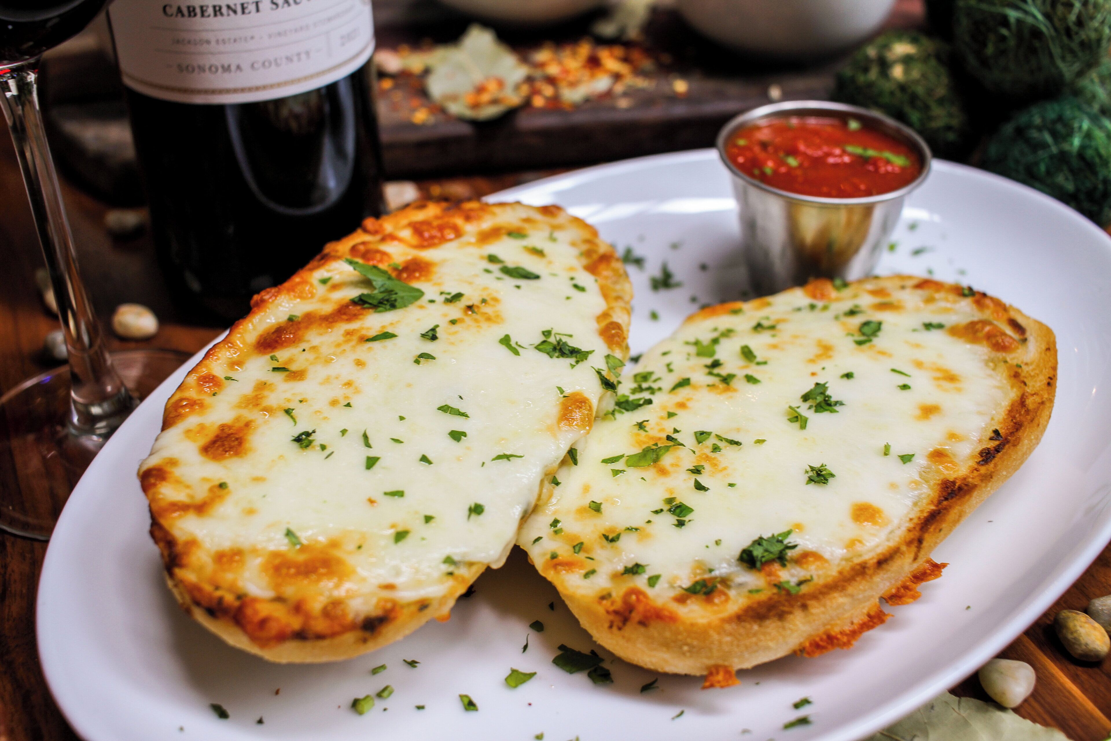 Une assiette de pain à l’ail, nappé de fromage fondu et parsemé d’herbes, une bouteille de vin et un petit bol de sauce marinara.