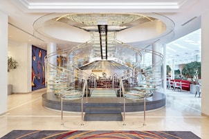 Un hall d’hôtel moderne doté d’un escalier central en verre, d’un espace salon avec des tables et des chaises, et d’un grand tableau abstrait accroché au mur.