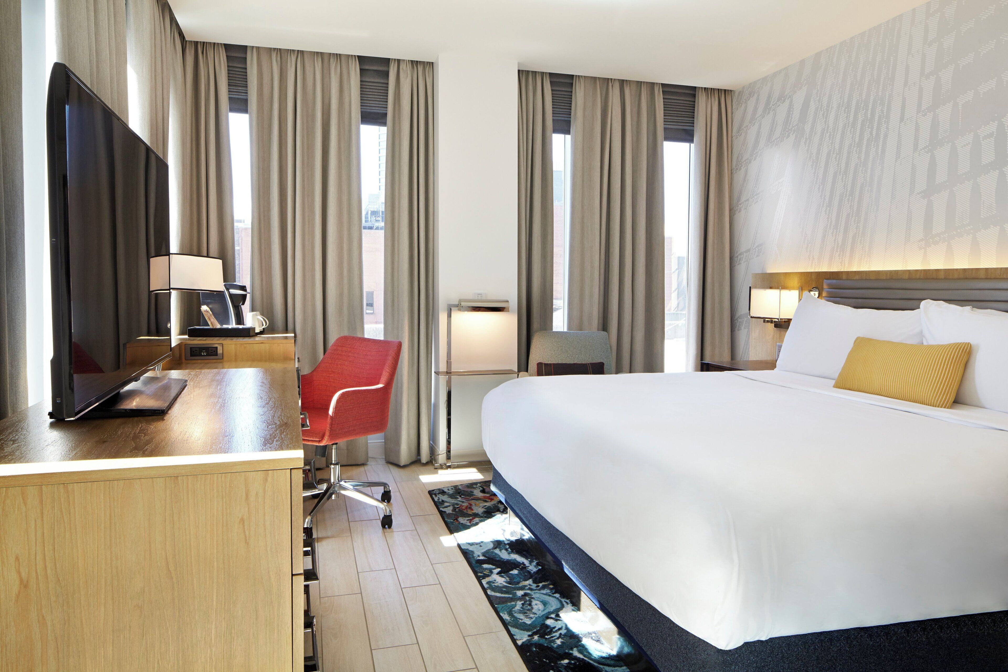A hotel room with a bed, a desk with a flat-screen TV, a red chair, a desk lamp, and a nightstand.