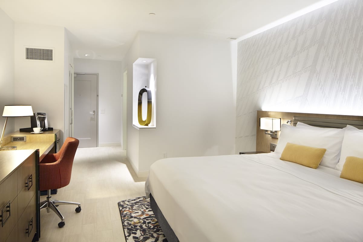 Habitación de hotel con cama, escritorio con silla, lámpara y estante en la pared.