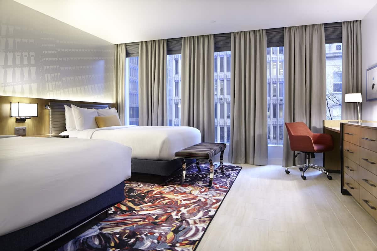 Habitación de hotel con dos camas, un escritorio, una silla, un ventanal con cortinas y vistas a la ciudad.