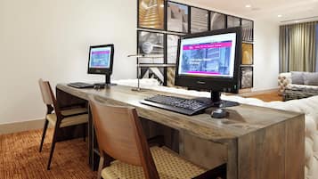 A hotel room with a long wooden desk, two monitors, a keyboard, and a mouse. There are two chairs with woven seats. The room has a large window with curtains.