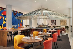 A modern restaurant with a central bar, round tables, and vibrant abstract art on the walls.