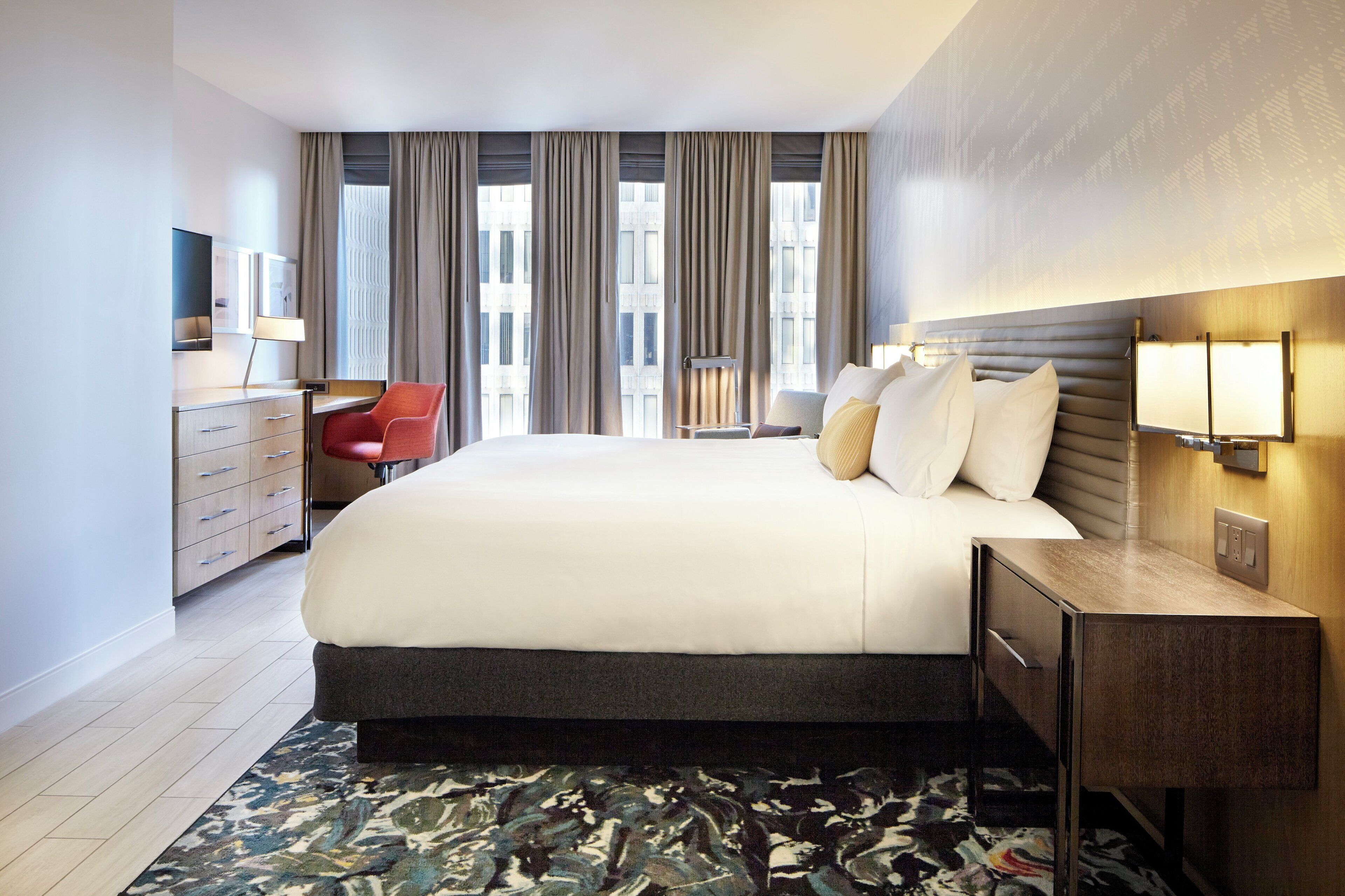 Habitación de hotel con una cama grande, un escritorio, una silla y vistas a la ciudad a través de la ventana.