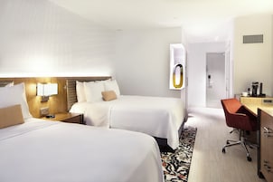 Une chambre d’hôtel avec deux lits, un bureau, une chaise, une machine à café et une fenêtre avec des stores.