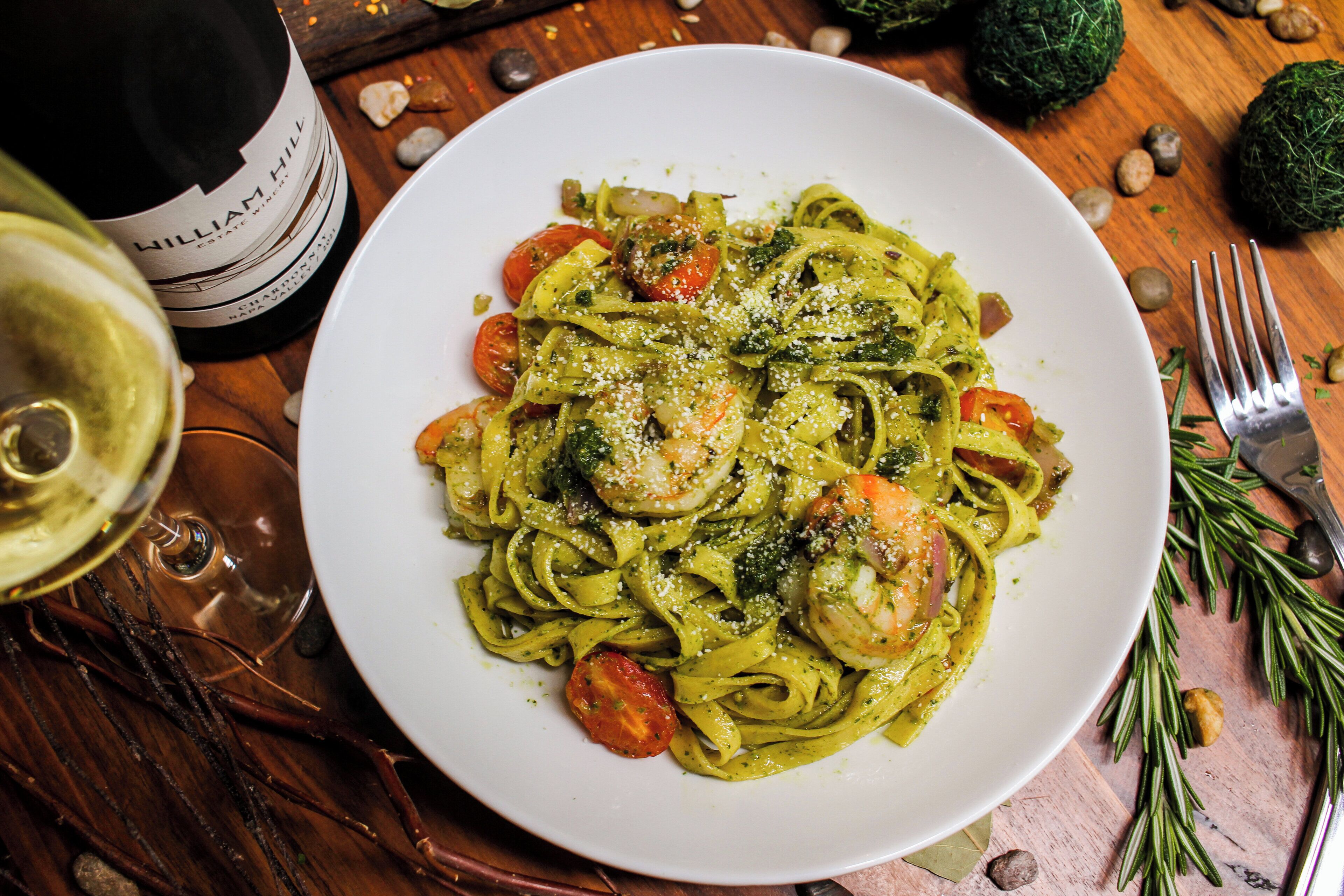 Een bord pasta met garnalen en groenten, een fles wijn en een vork op een houten tafel.
