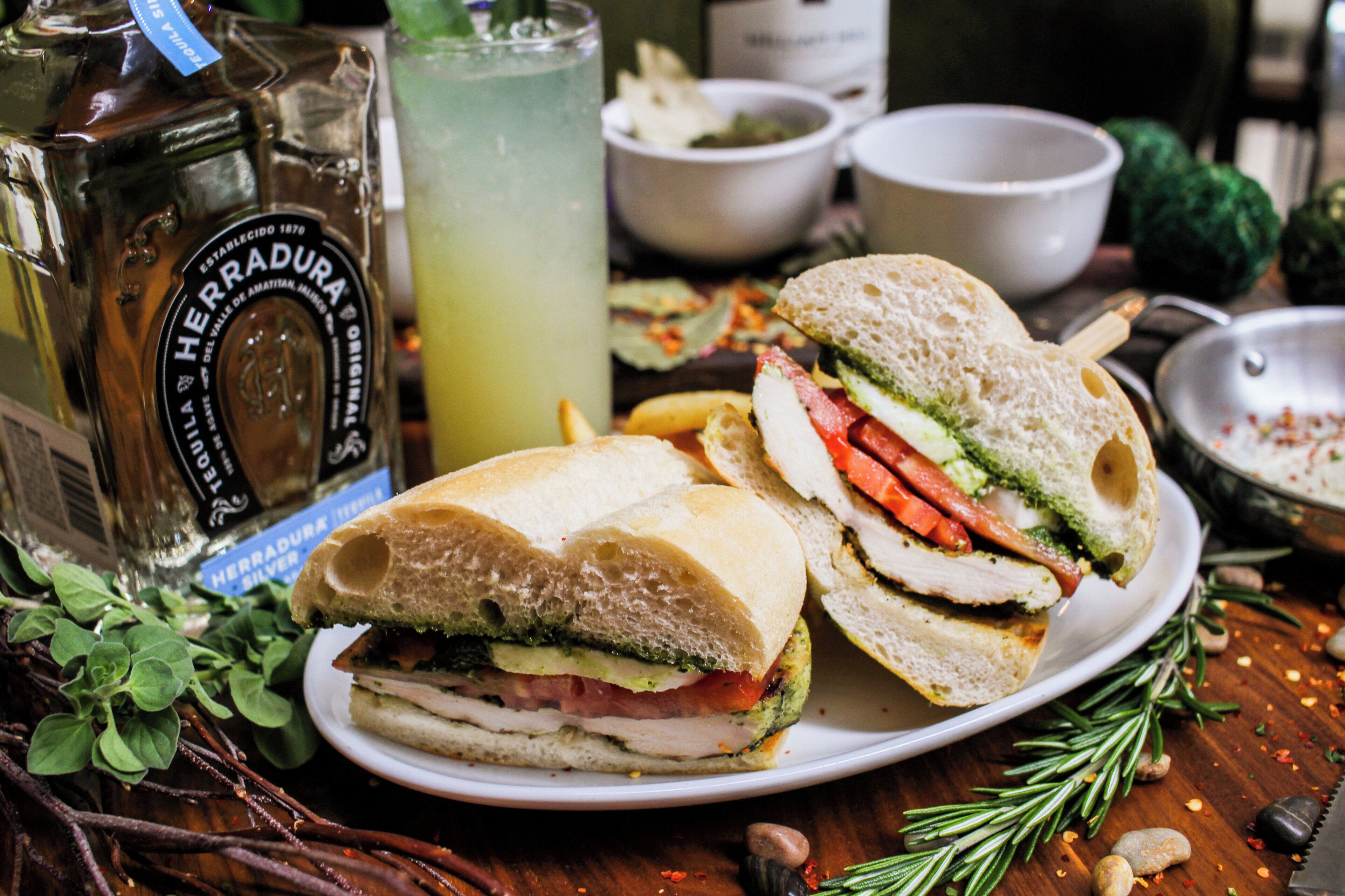 A sandwich with various fillings, a bottle of Herradura tequila, a glass of a green beverage, and some bowls and vegetables on a table.