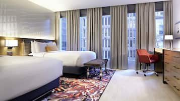 Habitación de hotel con dos camas, un escritorio, una silla y vista a la ciudad a través de amplias ventanas.