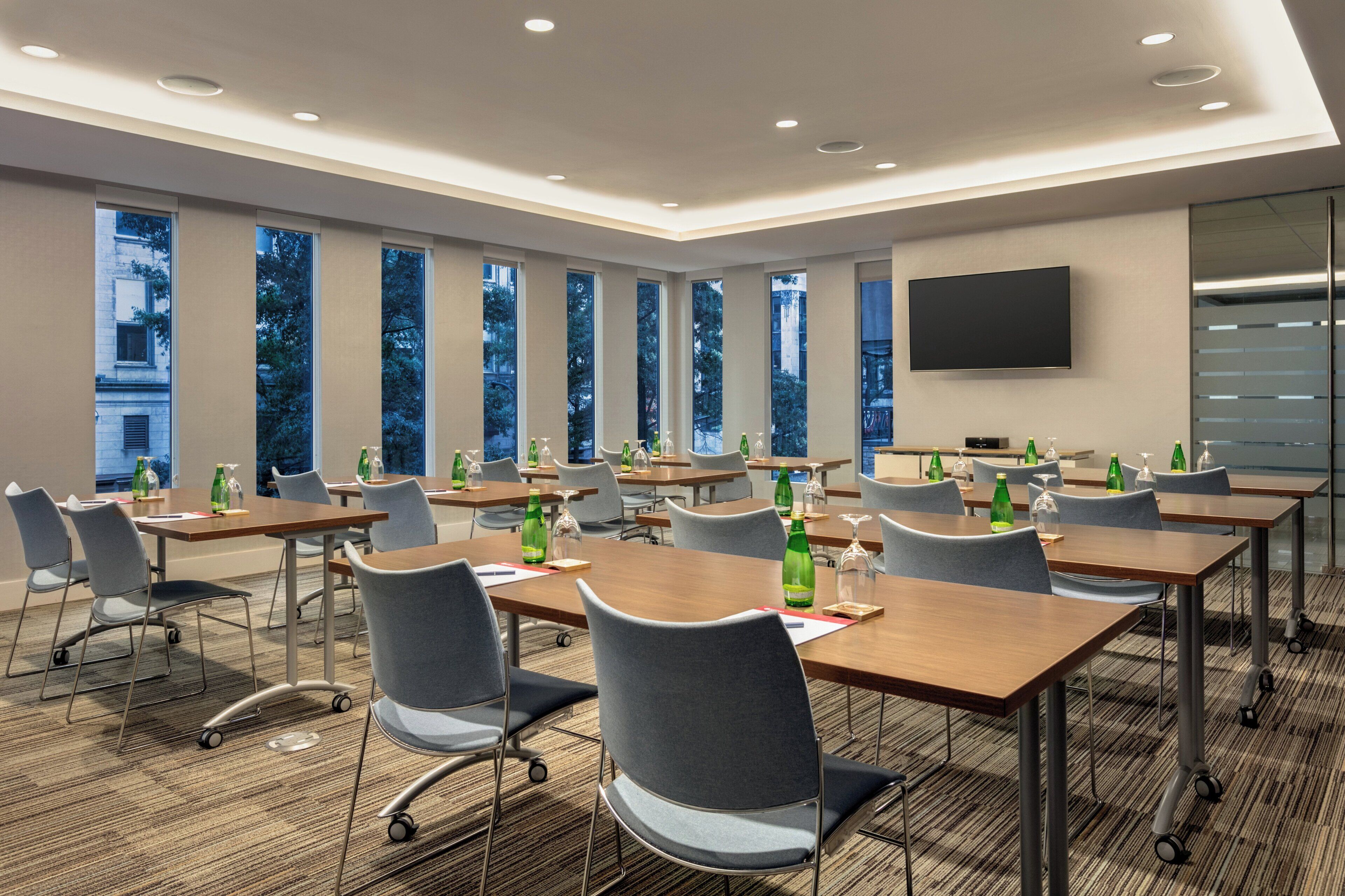 A conference room with multiple tables and chairs, large windows, and a flat-screen TV on the wall.